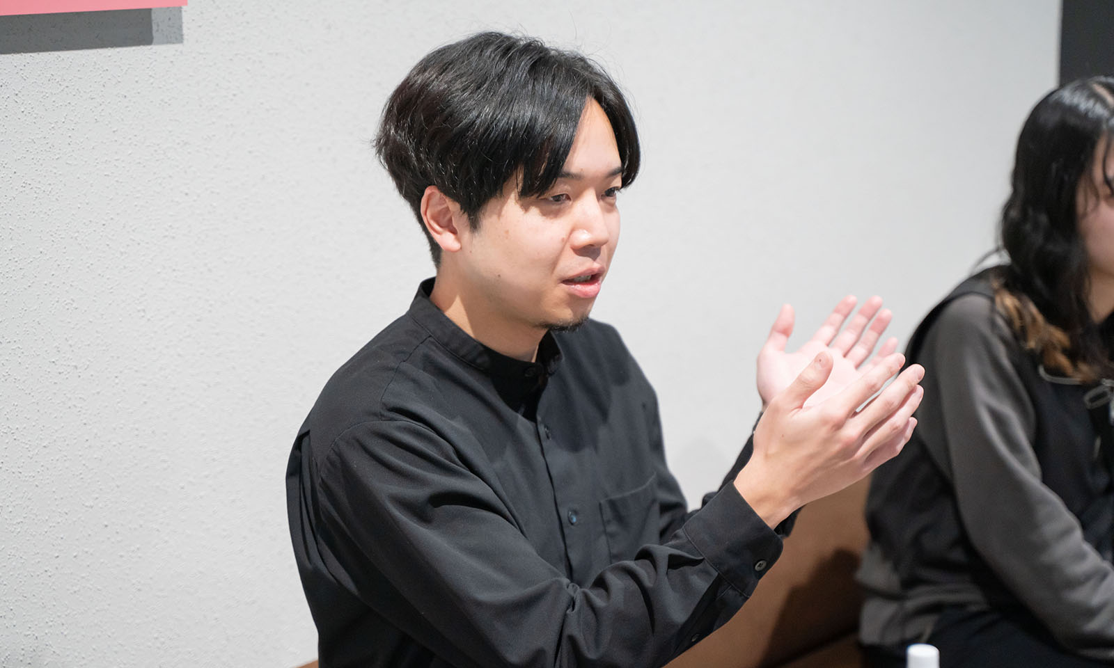 ALTはじまり。襟のない黒の長袖シャツを着る千田さんが手をろくろを回しているかのように広げて説明をする姿。耳まである髪の毛も黒、優しい中に強い眼差しをしている。ALTおわり。