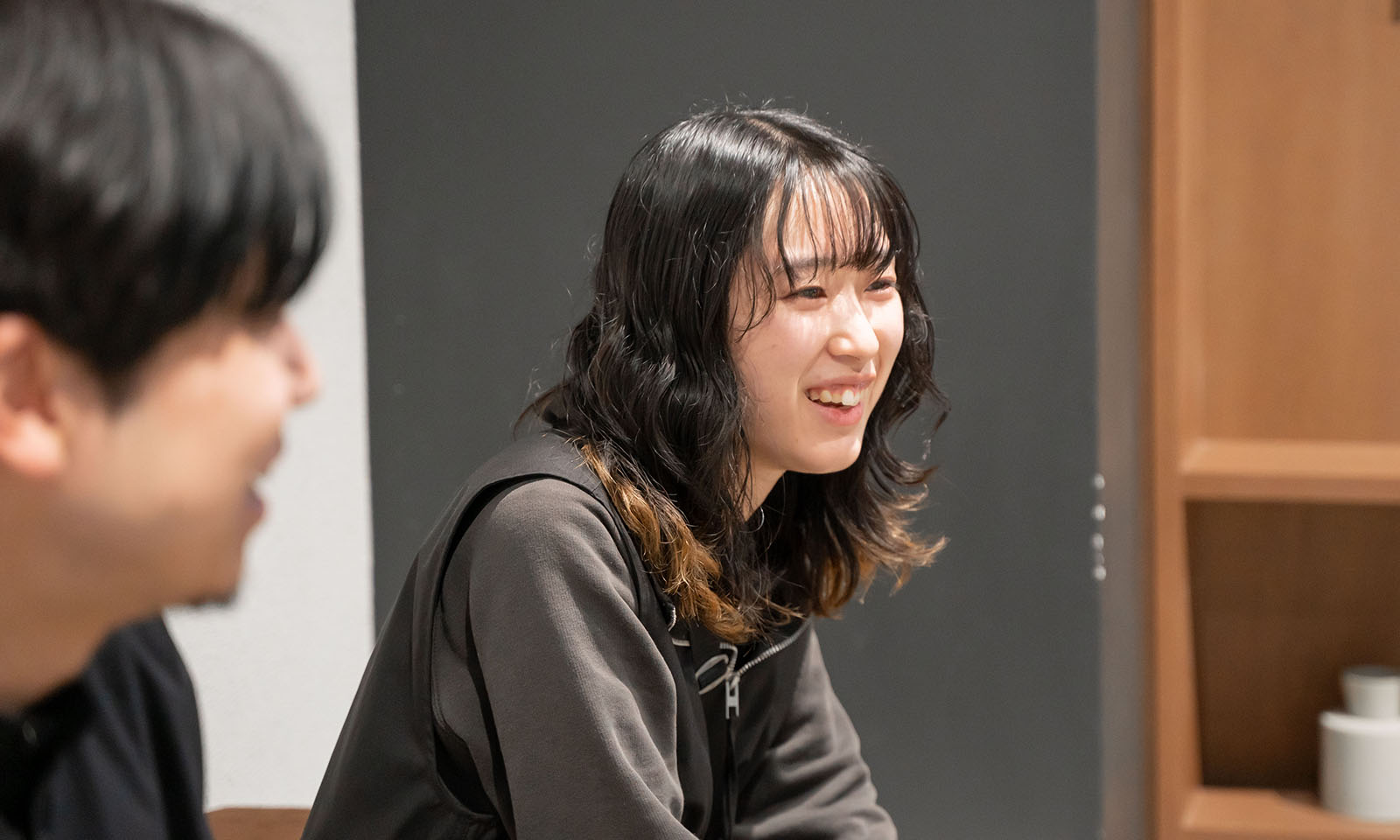 ALTはじまり。背景が木目とグレーのおしゃれなコクヨのオフィスで、櫻井さんが笑顔で話す様子。ウェーブがかかっている黒髪に、毛先が明るい茶色になっている。グレーのロンティに黒いベストを着ている。ALTおわり。