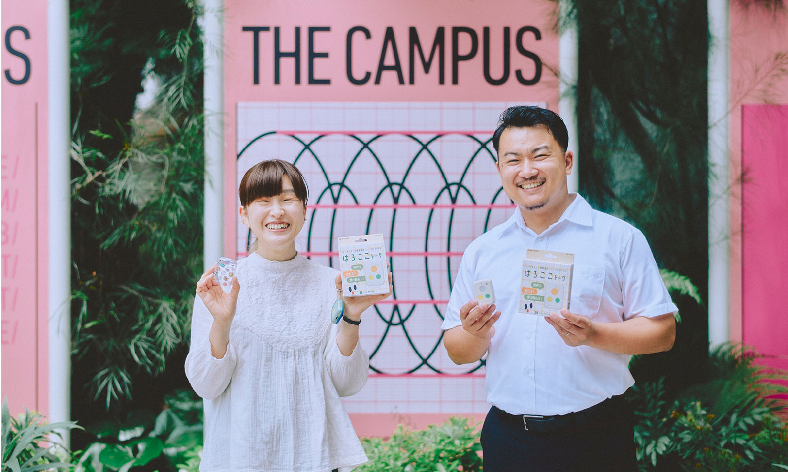 <ALT始まり>THE CAMPUSのピンク色の看板の前ではろここトークを手に並ぶインタビュイーの2人。トップスの白とボトムスの黒が揃っていて、素敵な笑顔を見せている。<ALT終わり>