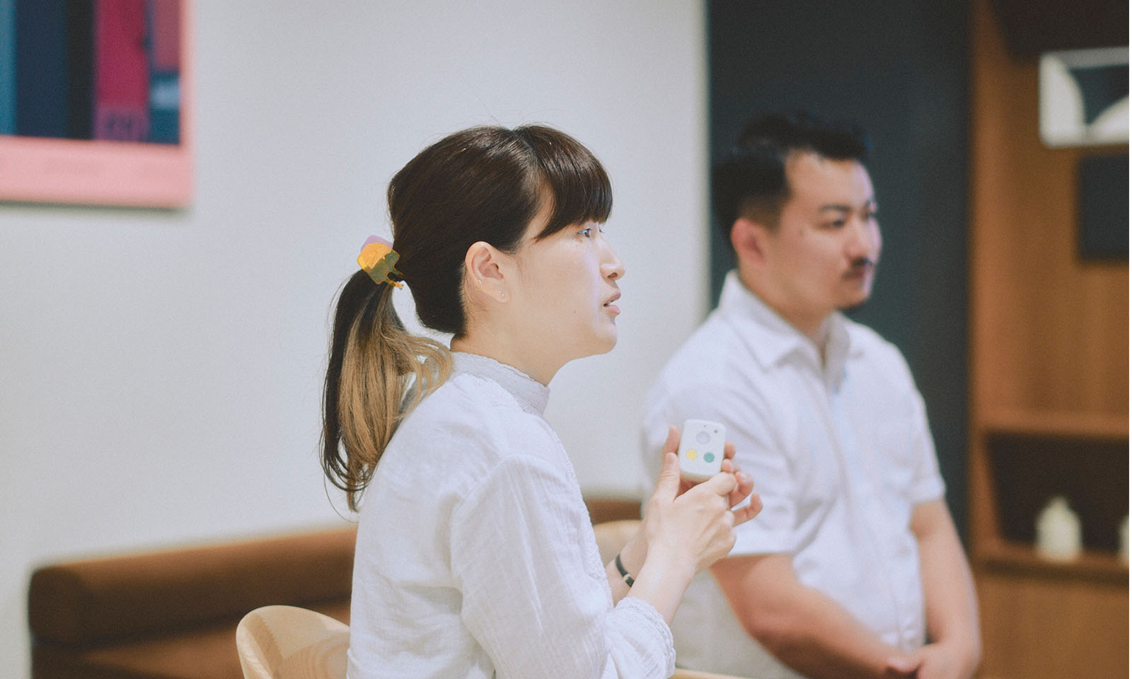 <ALT始まり>製品を手に、真剣な表情でインタビューに答える横田さんの横顔。金と黒のツートーンの背中程度の長い髪を耳の高さでポニーテールにしている。<ALT終わり>