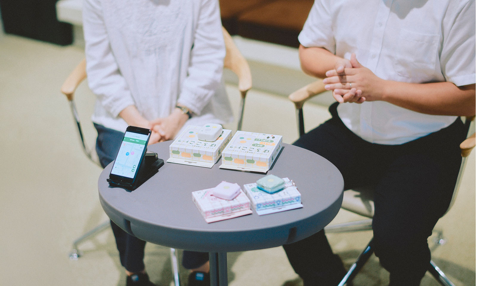 <ALT始まり>グレーの円形テーブルの上に並べられた旧タイプの製品とはろここトーク、位置情報を示すアプリを開いた状態のスマートフォンの写真。インタビュー中、これらを手にしながら話をしていただいた。<ALT終わり>