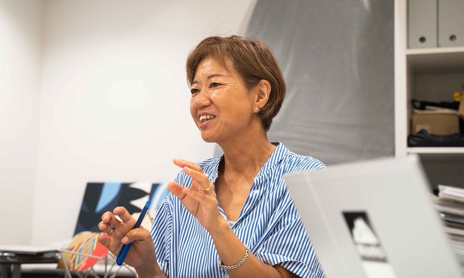 ALTはじまり。明るい茶色のボブヘアーを耳にかけ、少し日焼けした肌に爽やかな青のストライプワンピースを着ているコクヨのこうもりさん。手で何かを形作りながら、真剣な面持ちで、平井先生に語りかけている。ALTおわり。