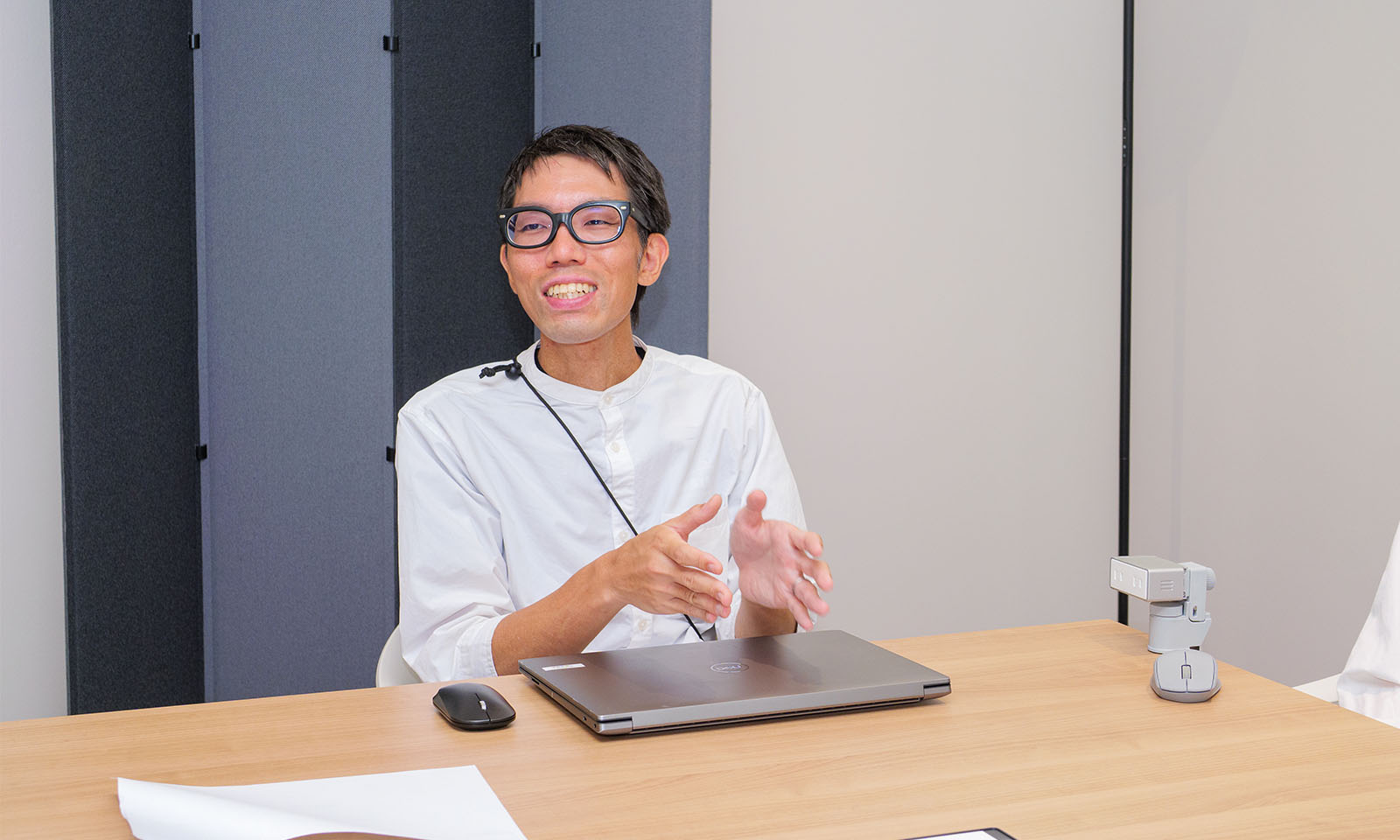 ALTはじまり。会議室に置かれたモノクロのパーティションをバックに、黒縁のメガネをかけた白シャツの男性が柔和な笑顔で語りかけている。手前には閉じた状態のノートパソコンが置かれ、その上で身振り手振りをしながら丁寧に説明をしている。ALTおわり。
