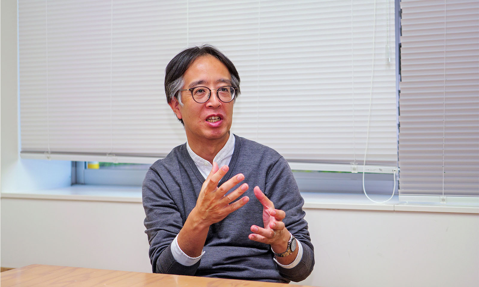 ALTはじまり。窓にかかるブラインドを背に、メガネをかけた白髪まじりの男性が両手で身振り手振りをしながら喋っている。襟のない白シャツに長そでのグレーのカットソーを重ねた装いで、左腕にはシルバーの腕時計を付けている。ALTおわり。