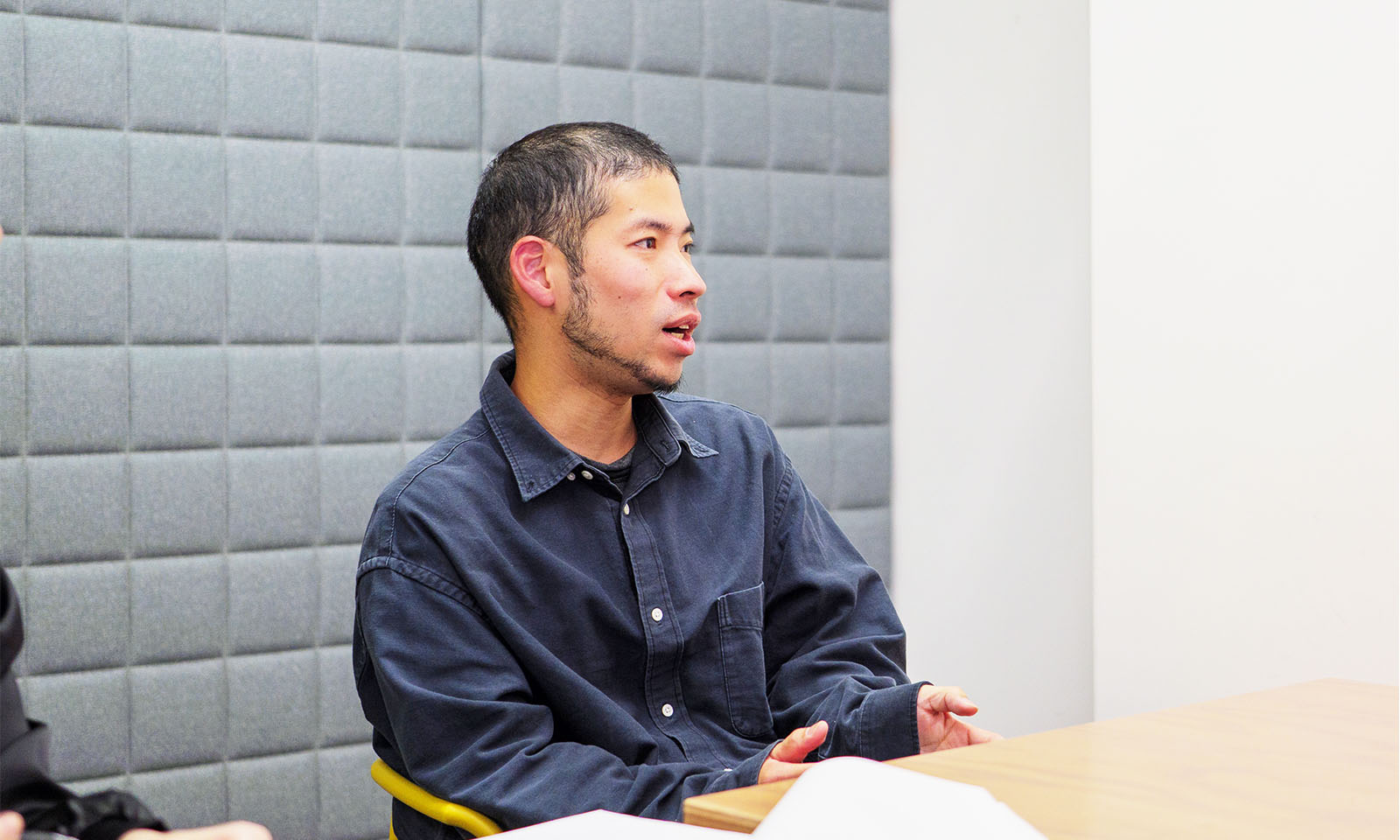 ALTはじまり。クッションタイプのグレーのパーティションが背後にある空間。耳が見えるほど短く切りそろえた髪と、もみあげまでつながった濃い髭が印象的な男性が、左に目線を向けながら喋っている。襟のあるネイビーの長そでを着用。ALTおわり。