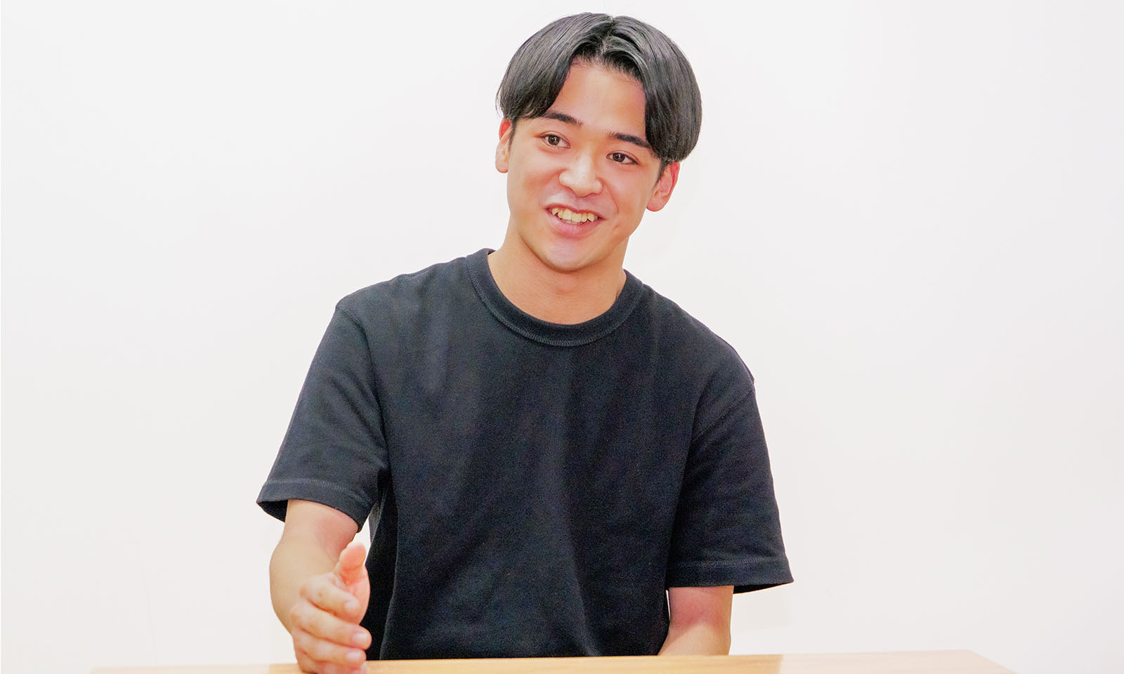 ALTはじまり。白い壁が広がるシンプルな空間。前髪をセンターで分けた短髪の男性が、笑顔を含みながら開いた右手をテーブルの上に立てて喋っている。襟の丸い黒のTシャツを着用。ALTおわり。