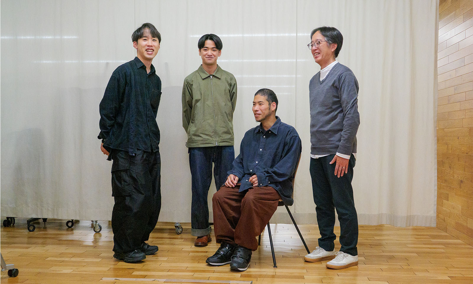 ALTはじまり。白色のカーテンをバックに4名の男性が並び、カメラに向かって優しく微笑みかけている。右から2番目の男性は、開発したばかりの新しいイスに深く腰かけ、少し斜めを向いて座っている。ALTおわり。