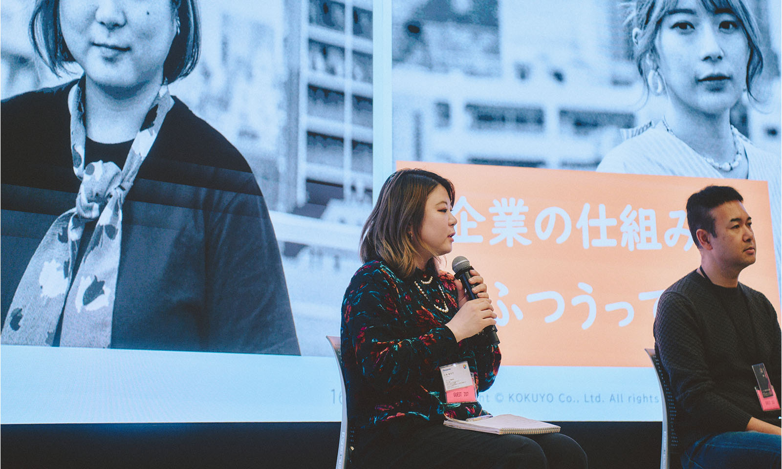 （ALT始まり）登壇者の1人、しらとりまいこさんが椅子に座り、スクリーンの前でマイクを持って話す様子。肩に触れるくらいのボブヘアは暗い茶髪がベースで、襟足にかけて金髪のグラデーションがかかっている。黒地に赤や青や緑のカラフルな模様が華やかなトップスに、パールのアクセサリーを重ねている。（ALT終わり）