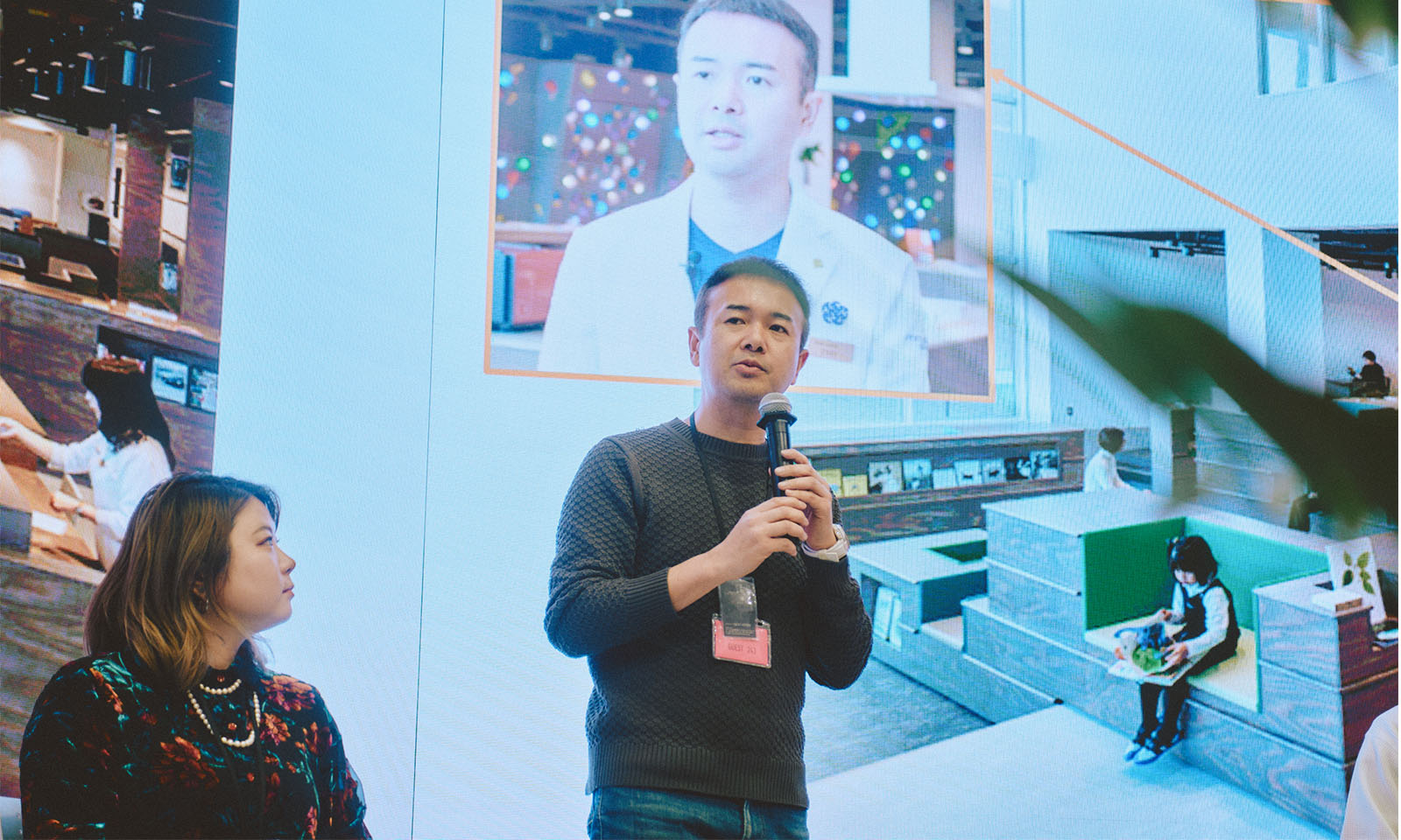 （ALT始まり）登壇者の1人、みやけたくさんがスクリーンの前に立ち、マイクを持って話す様子。後ろのスクリーンには白衣姿のみやけさんが映っている。グレイヘアのすっきりとした短髪、落ち着いたグレーのニットにデニムという服装。（ALT終わり）