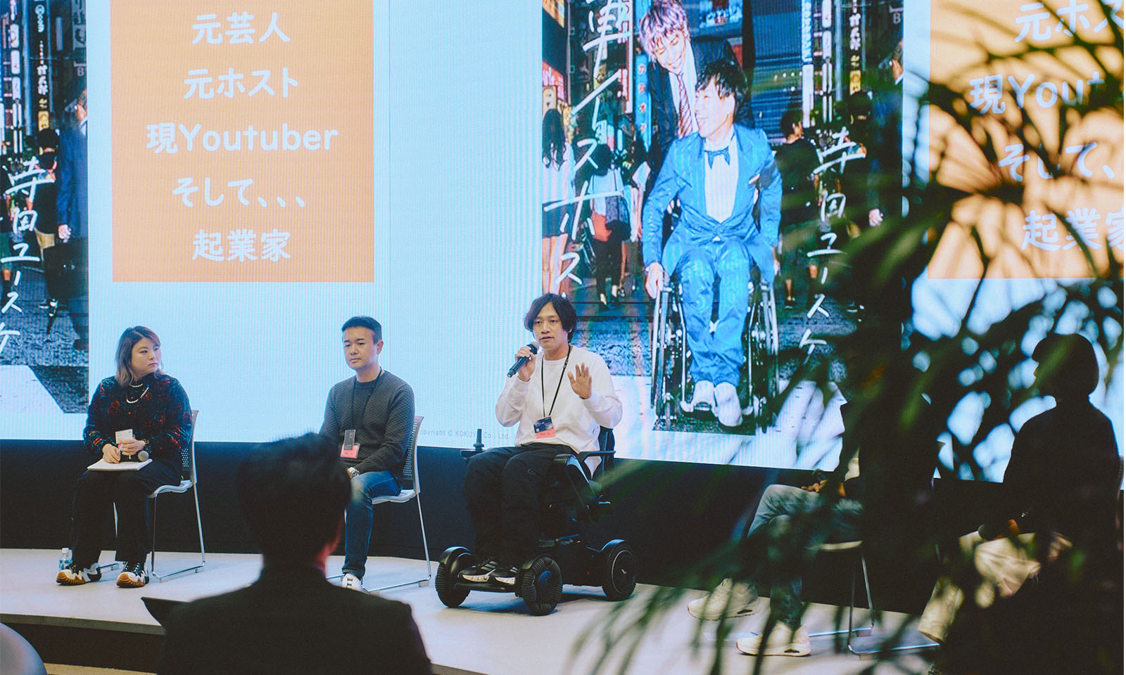 （ALT始まり）登壇者の1人、てらだユースケさんが電動車椅子に座りマイクを持って話す様子。白鳥さん、三宅さんが同じ壇上でてらださんの話を聞いている様子が一枚に写されている。長めの前髪をセンターで分け、白いトップスに黒のパンツとスニーカーでカジュアルな服装。（ALT終わり）