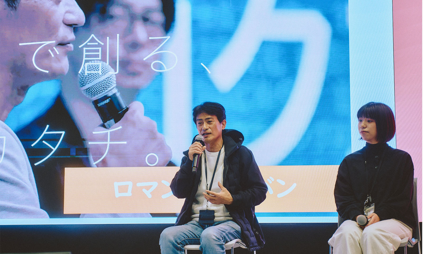 （ALT始まり）登壇者の1人、みやざわのりともさんが椅子に座りマイクを持って話す様子。白いカットソーに紺色のパーカーを羽織り、デニムパンツを合わせているカジュアルな服装。隣には同じく登壇者のたかぎさんが話を聞いている様子も写っている一枚。（ALT終わり）