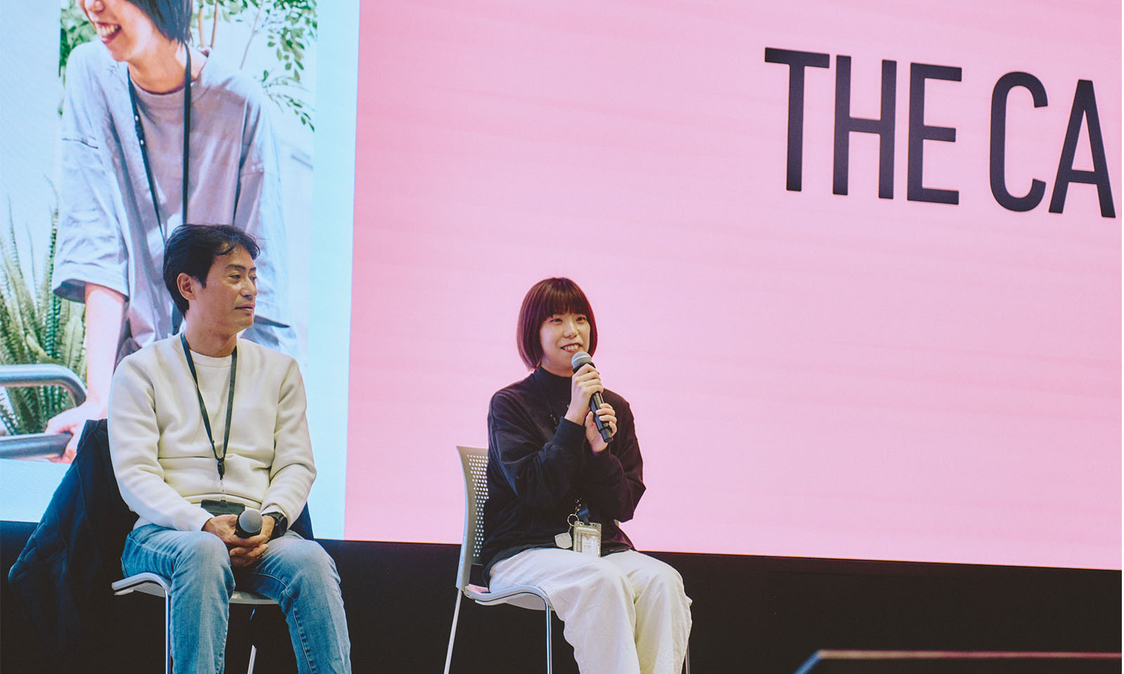 （ALT始まり）登壇者の1人、たかぎりほさんがスクリーンの前で椅子に座り、マイクを持って話す様子。ストレートのショートボブに黒色のニット、白いパンツを合わせた服装で、微笑みながら話をしている。隣には同じく登壇者のみやざわさんが上着を脱いだ状態で話を聞いている様子が写っている。（ALT終わり）