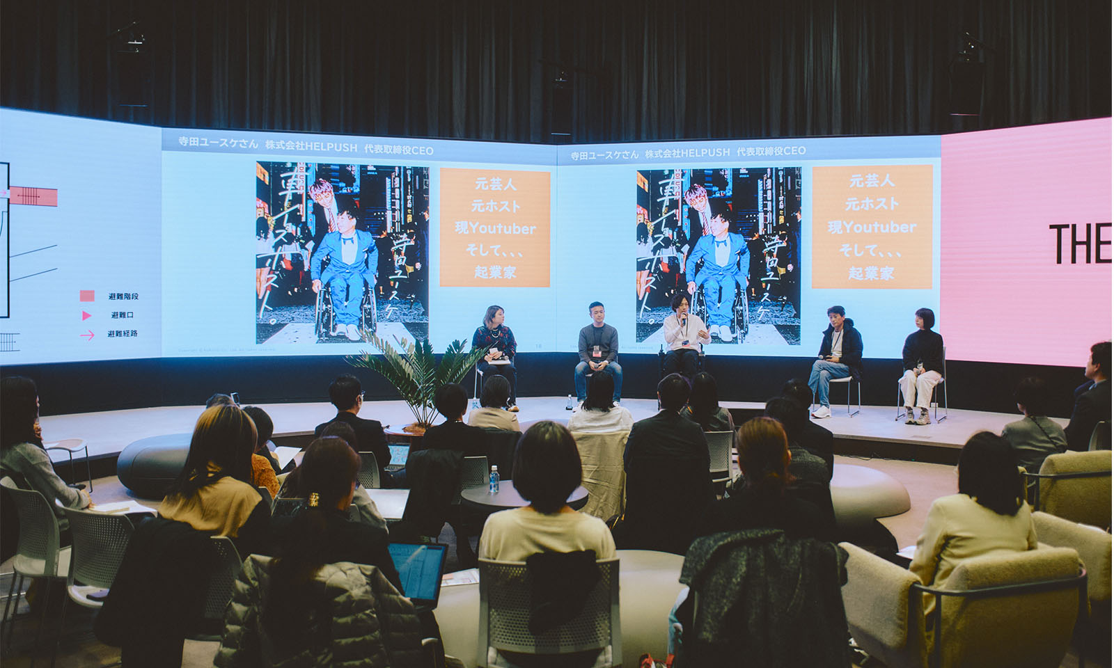 （ALT始まり）会場全体の様子。前にある大きなスクリーンは扇形になっていて、どの席からも見えやすい。参加者の座る椅子もソファや椅子など多様になっている。（ALT終わり）