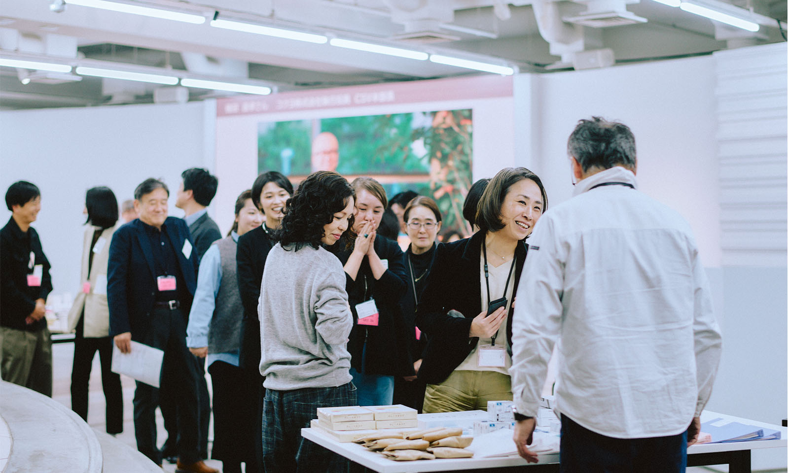 （ALT始まり）帰り際、お土産を受け取るため列になって並びながら会話を楽しむ参加者たち。打ち解けた雰囲気でリラックスしている表情が見える。（ALT終わり）