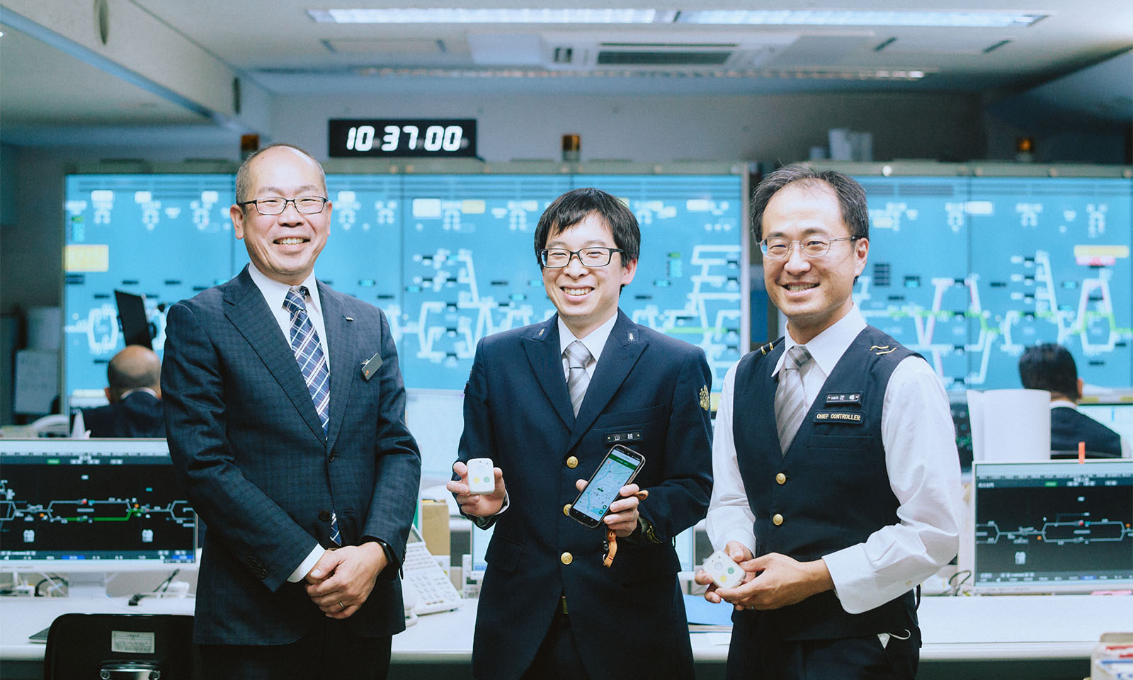 （ALT始まり）今回取材をおこなったJR東海の3名。それぞれスーツと制服を着用し、はろここトーク本体とスマートフォンを手に、＊＊でにこやかに笑っている。（ALT終わり）