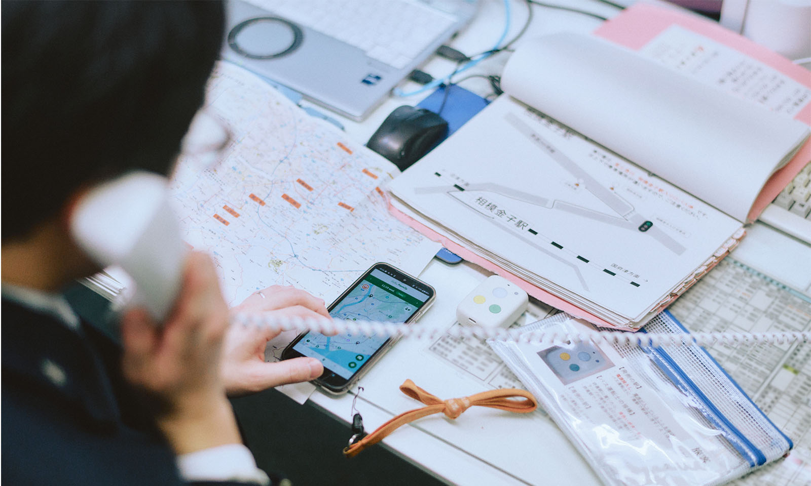（ALT始まり）デスク上で、電話をしながら端末のGPS機能を使う様子。路線図などの運行管理に必要なものも広げられている。（ALT終わり）