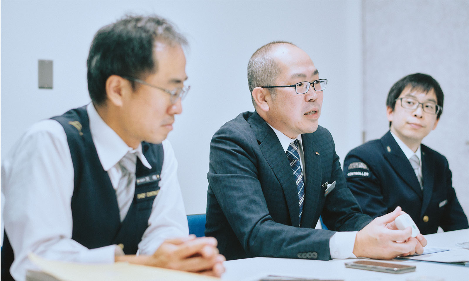 （ALT始まり）インタビューを受ける3名の様子。はろここトークの端末を手に語る向久保さんに視線を向けながらしっかりと耳を傾ける山越さんと花嶋さんのほか、机の上には分厚いファイルなどの資料も見える。（ALT終わり）