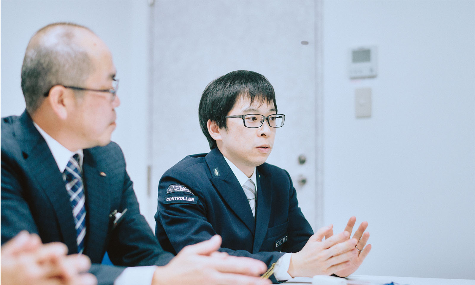 （ALT始まり）紺色ジャケットに白のシャツ、グレーのネクタイの制服を着用し、手振りを添えながら話す山越さん。（ALT終わり）