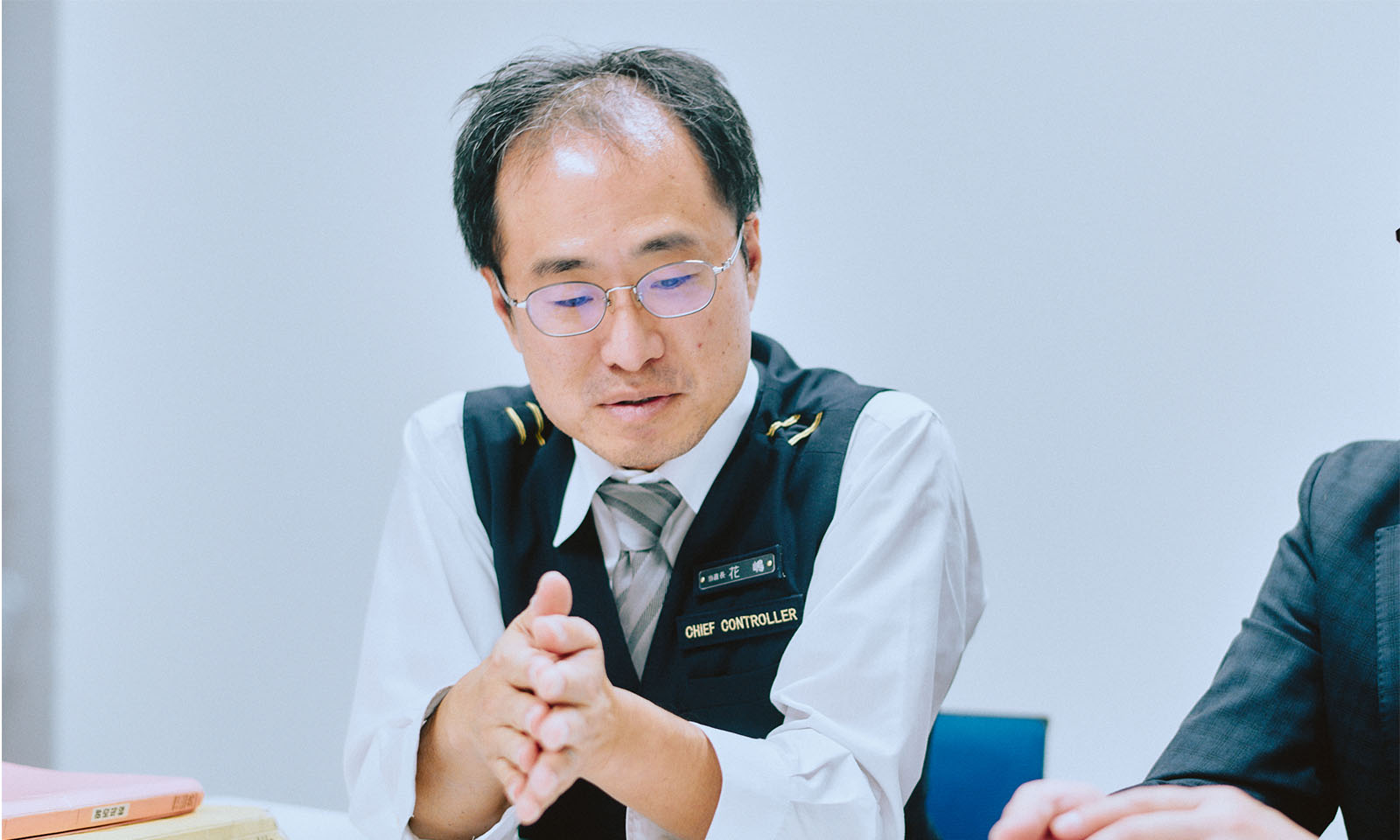 （ALT始まり）インタビューを受ける花嶋さん。紺色のベストに白い長袖シャツ、グレーのネクタイを着用し、落ち着いた表情で話をしている。（ALT終わり）