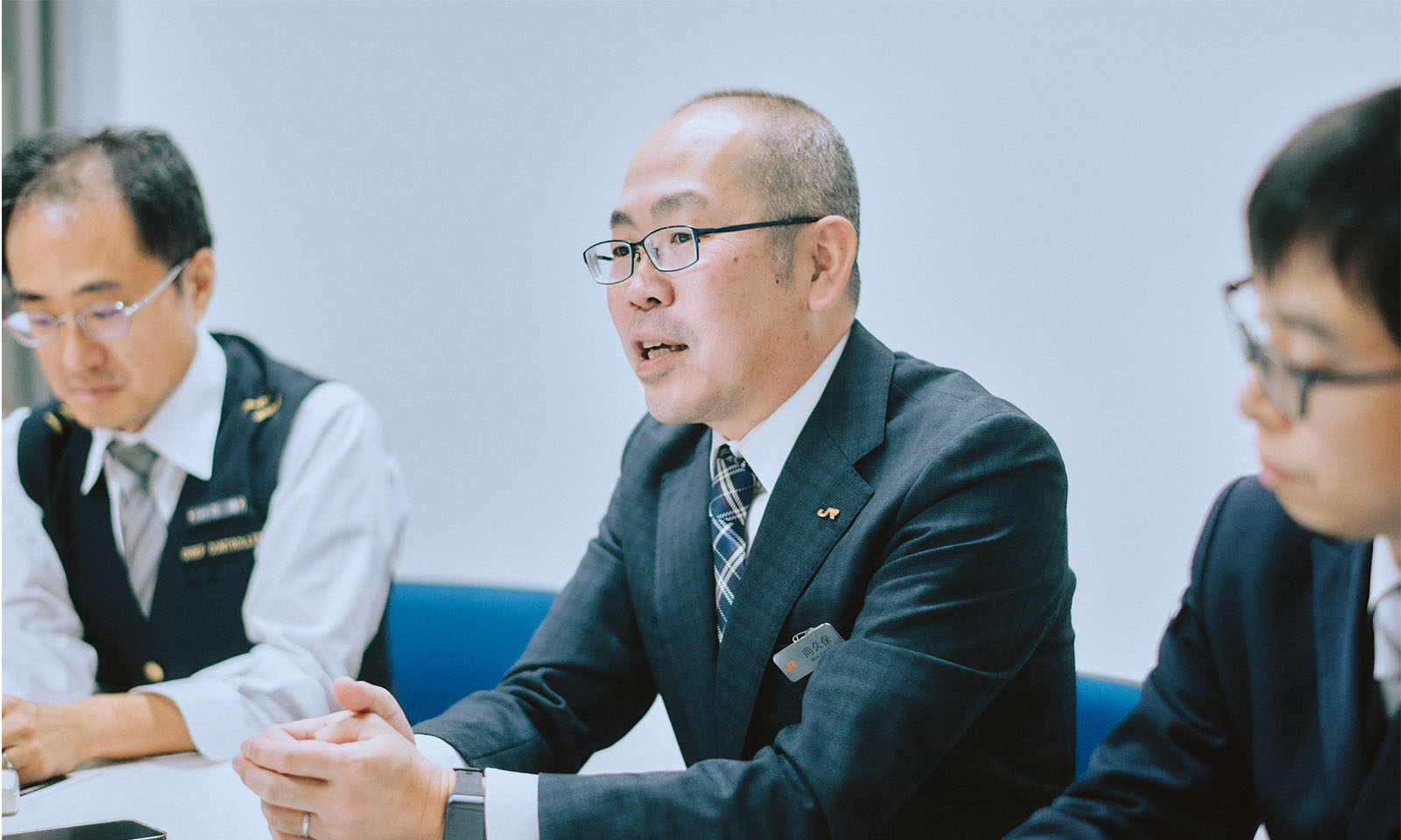 （ALT始まり）話をする向久保さん。落ち着いたチェック柄のネイビーのスーツを身に纏い、胸元にはJRのバッジが付けられている。（ALT終わり）