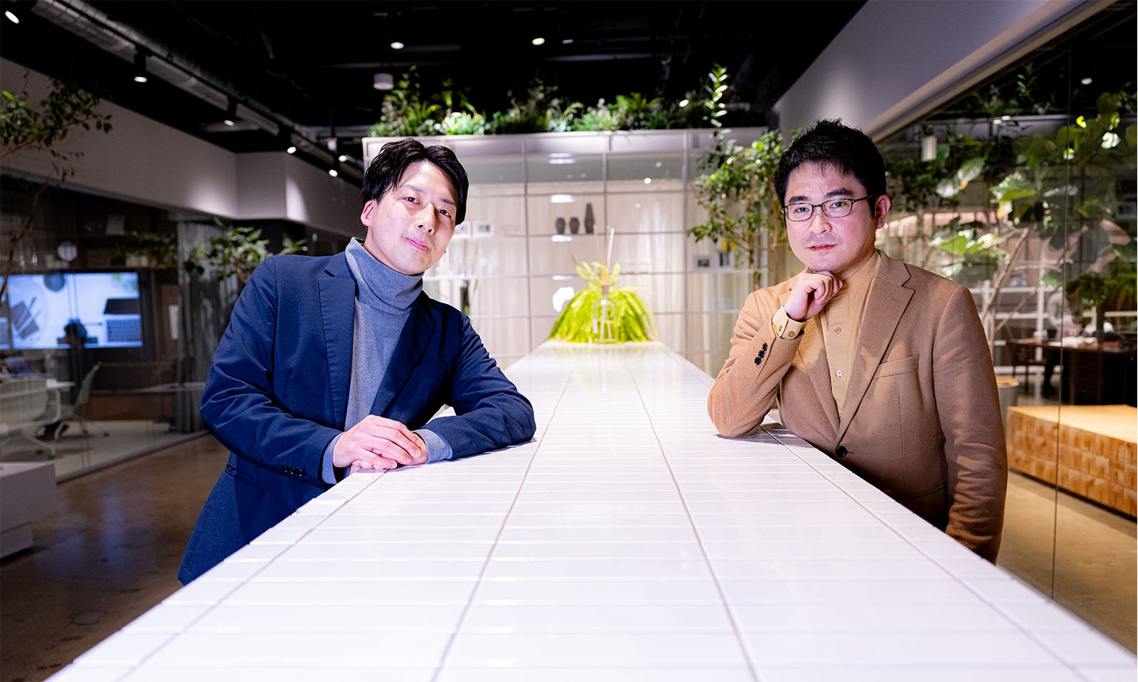 <ALT始まり>インタビュイーの紺色のジャケットを着た西村さんとベージュ色のジャケットを着た佐藤さんが、白いカウンターを挟んで立っている<ALT終わり>