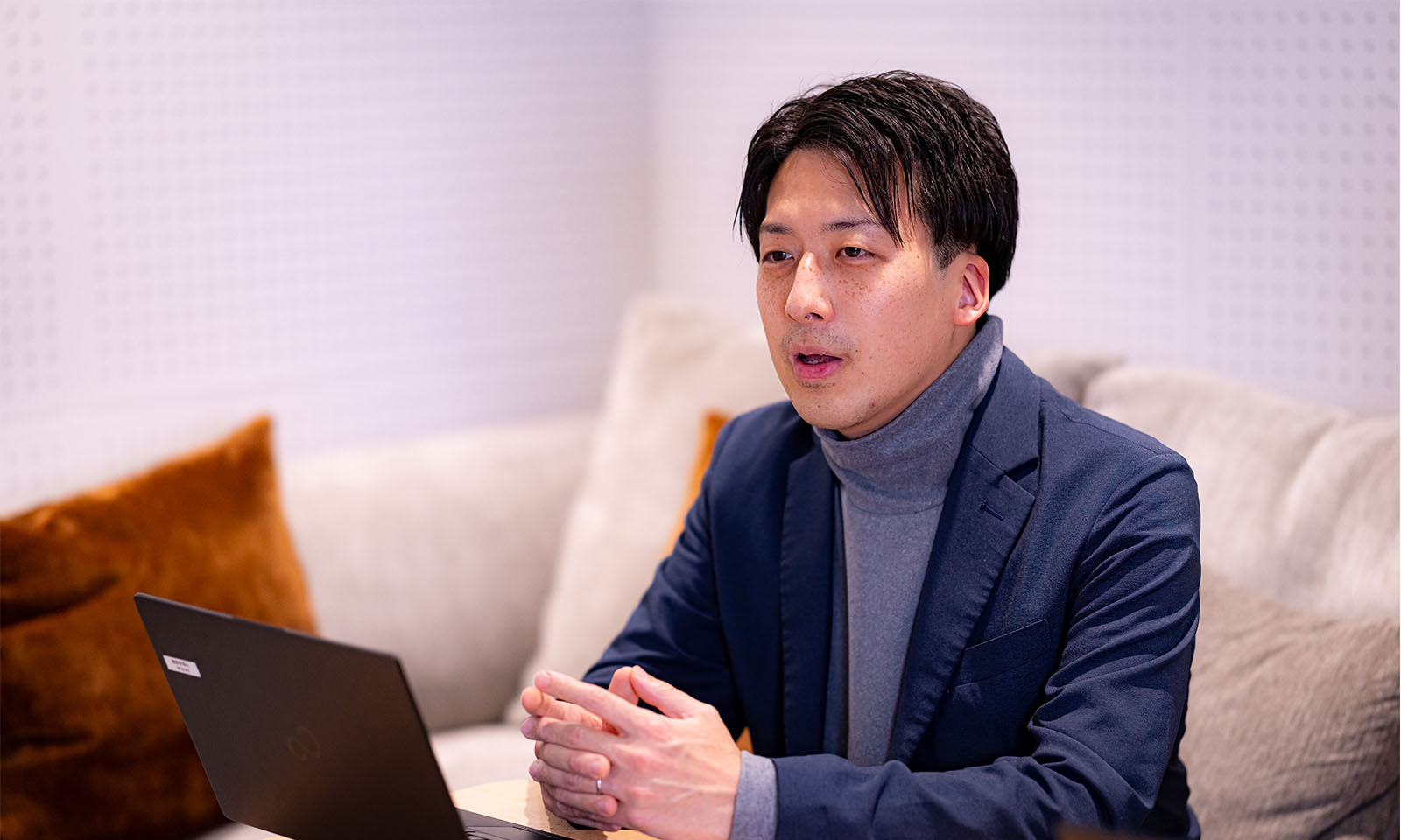 <ALT始まり>ソファに座っている紺色のジャケットを着た西村さん。テーブルの上のパソコンの手前で両手を組んでいる。<ALT終わり>