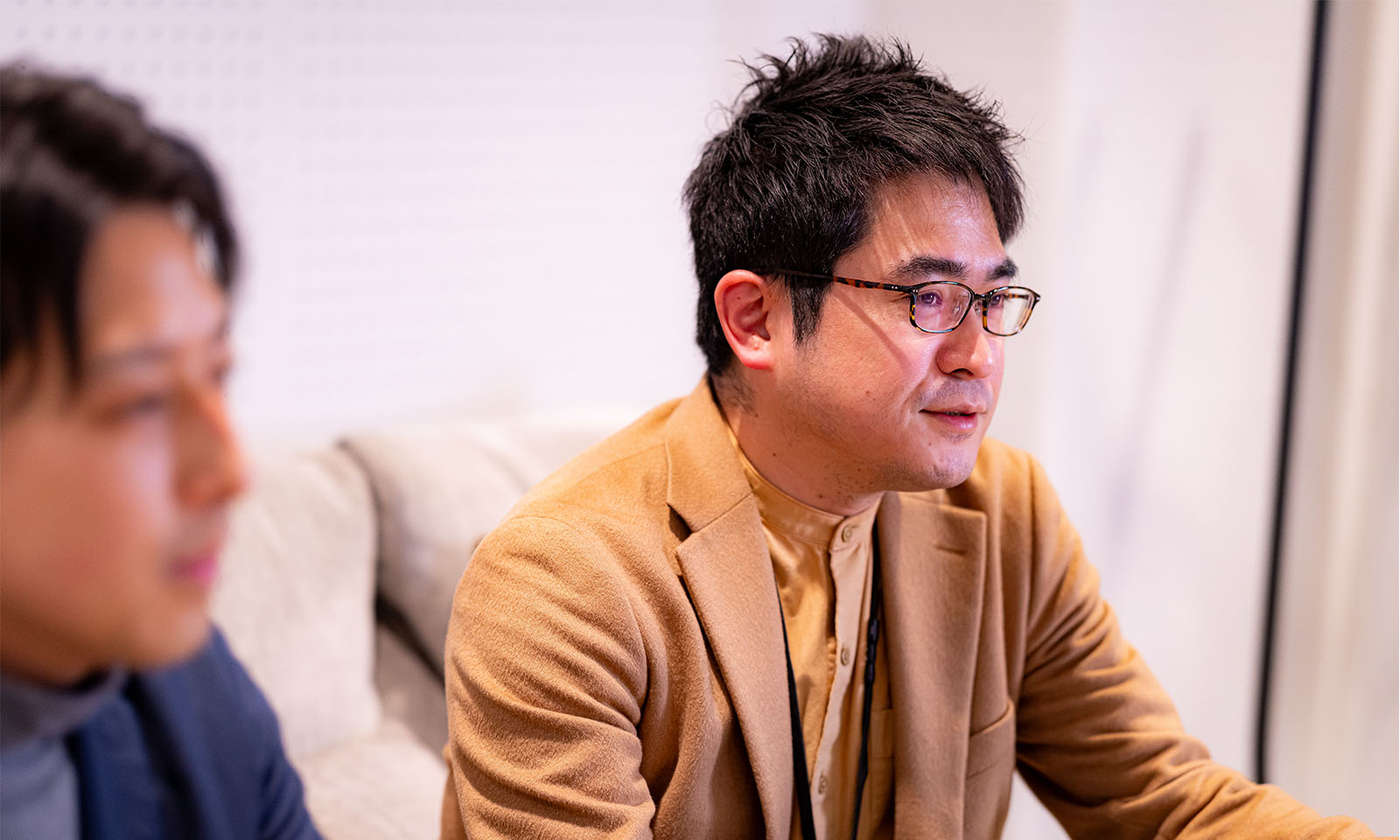 <ALT始まり>ベージュ色のジャケットを着て、眼鏡をかけている佐藤さんが、質問に対し、手振りを交えて回答している。<ALT終わり>