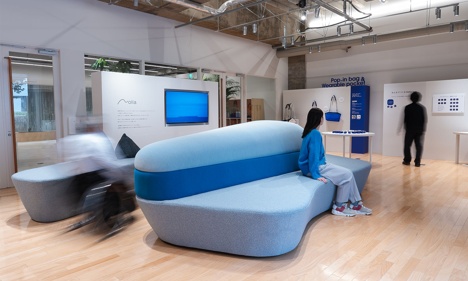 （ALT始まり）ブルーのソファが飾られた会場の様子。ソファの座面は海に浮かぶ島のような流線型で、ソファと同じ色味のニットを着た女性が腰掛けている。会場は白とブルーを基調としたさわやかな雰囲気で、壁に青いカバンなどが展示してある。（ALT終わり）