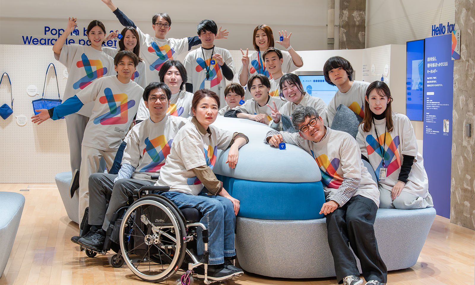 （ALT始まり）ブルーを基調としたソファを囲む、おそろいのTシャツを着た20人ぐらいの人々。手前には車いすに乗った女性と、背もたれに身体を預けてくつろぐ男性。後ろの列の男性は大きく万歳のポーズをとっている。（ALT終わり）