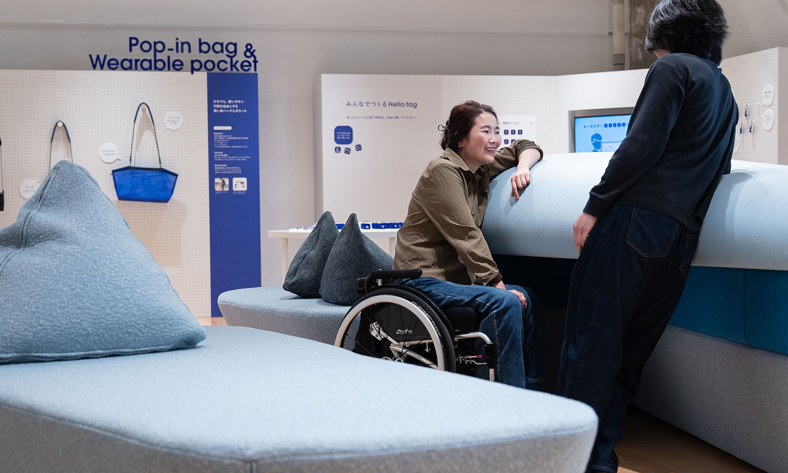 （ALT始まり）ソファの座面のくぼんだ部分に車いすを乗り入れて、背もたれに身体を預けてくつろぐ茶色いジャケットを着た女性。同じ背もたれにもたれて立つ黒い服の男性と話している。（ALT終わり）