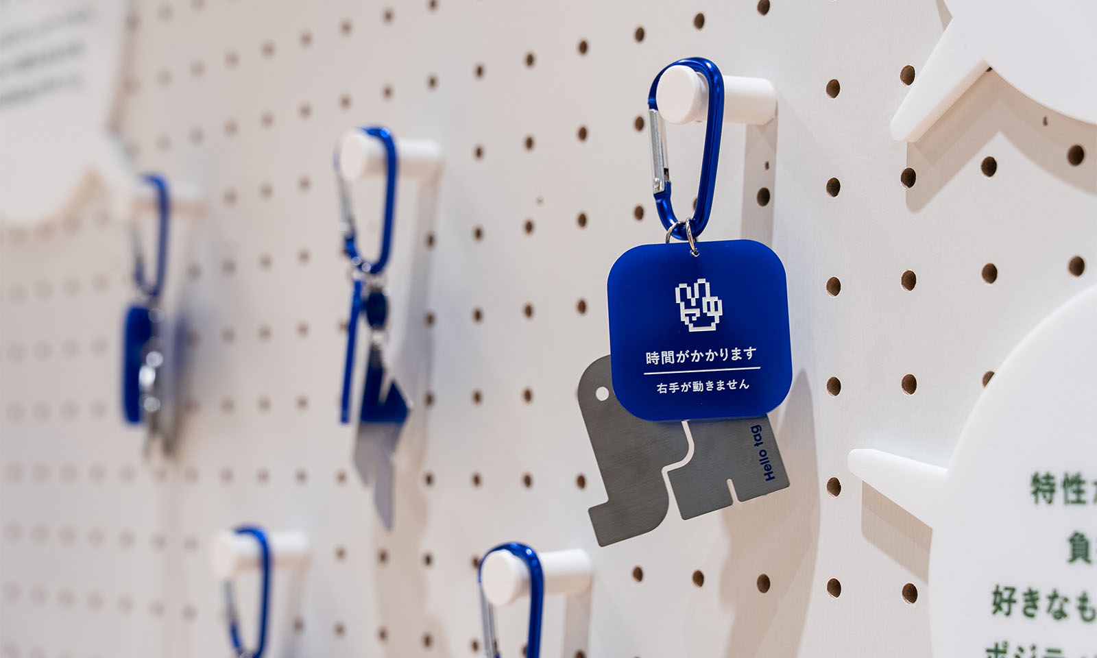 （ALT始まり）壁に、青いプレートのついたキーホルダーのチャームが吊り下げてある。チャームには「時間がかかります　右手が使えません」と白で印字してあり、ピースマークのイラストも描かれている。（ALT終わり）