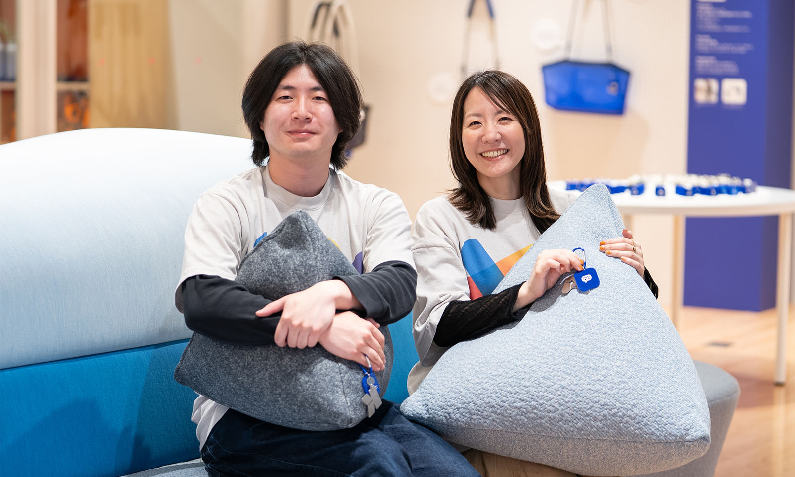 （ALT始まり）ブルーを基調としたソファに、おそろいのTシャツを着た男性と女性が並んで座っている。二人とも柔らかく微笑み、三角形のクッションを持っている。女性は片手で青いチャームを持っている。（ALT終わり）