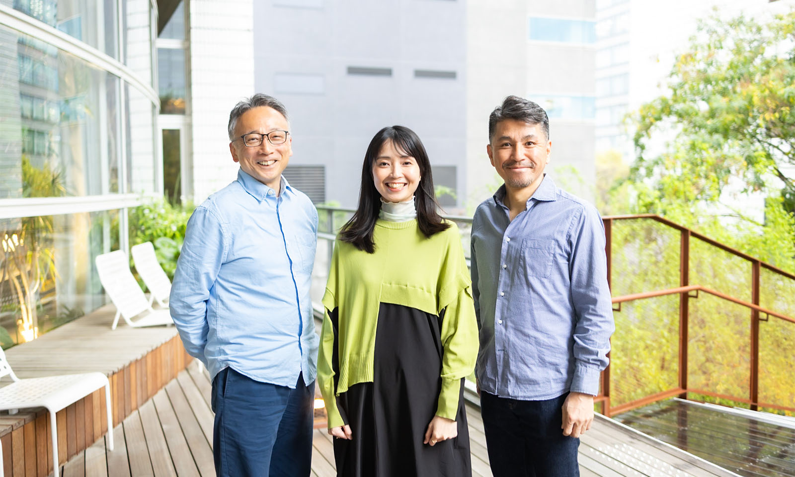 ALTはじまり。3人の人物が屋外に並んで立ってこちらを見ている。真ん中がグリーンのトップスで黒いスカートの女性、左が薄い水色のシャツの男性。右がブルーグレーのシャツの男性。ALTおわり。