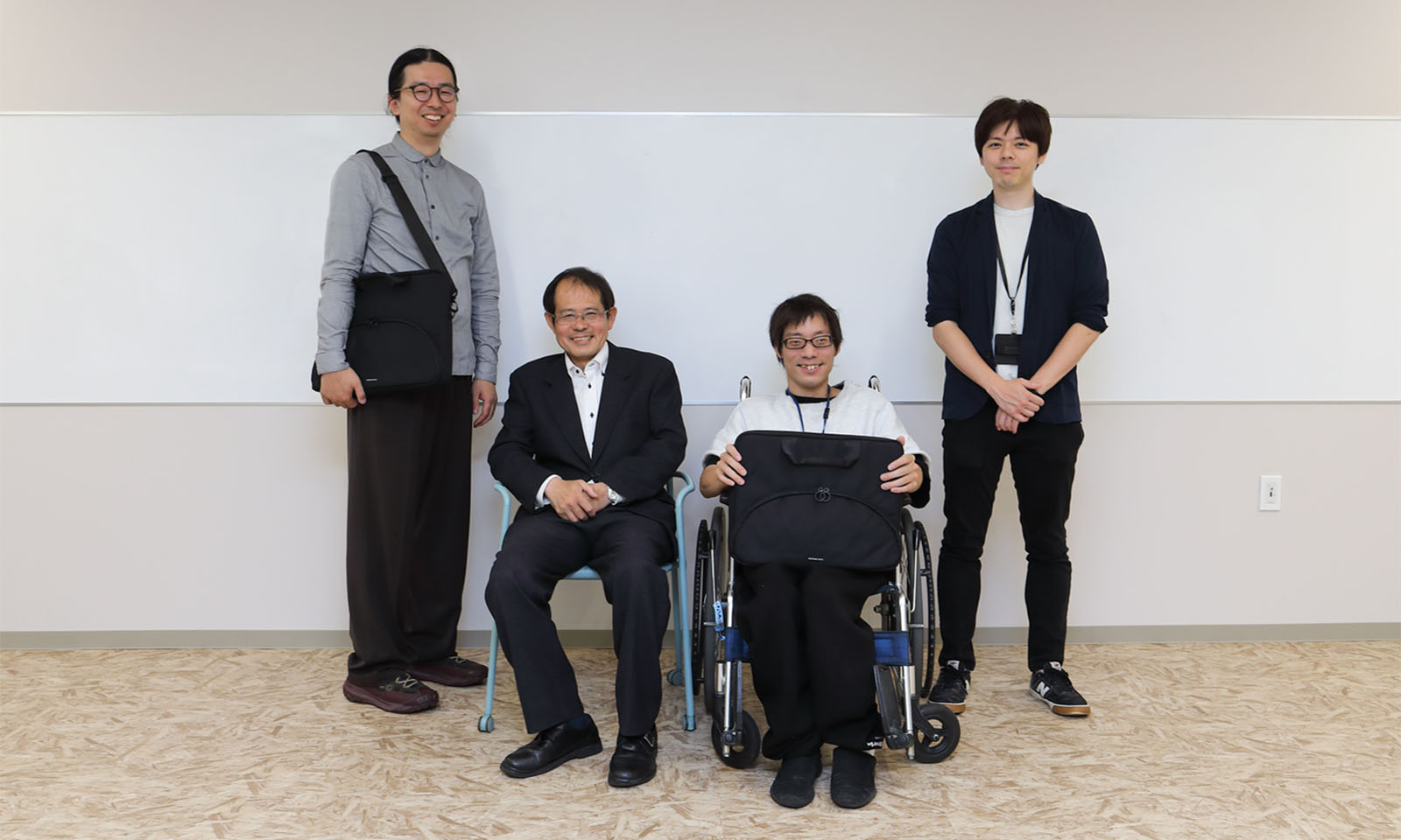 <ALT始まり>ホワイトボードの前に並ぶ男性4人の集合写真。左から、グレーのシャツを着て「耐衝撃パソコンバッグ 出し入れスムーズ前面ポケット」を肩に掛けた濃野さん、スーツ姿で椅子に座る中尾さん、車椅子に乗り膝に「耐衝撃パソコンバッグ 出し入れスムーズ前面ポケット」を置いた近藤さん、紺のジャケットを着た荒木さん。全員がカメラに向かって笑顔を見せている。<ALT終わり>