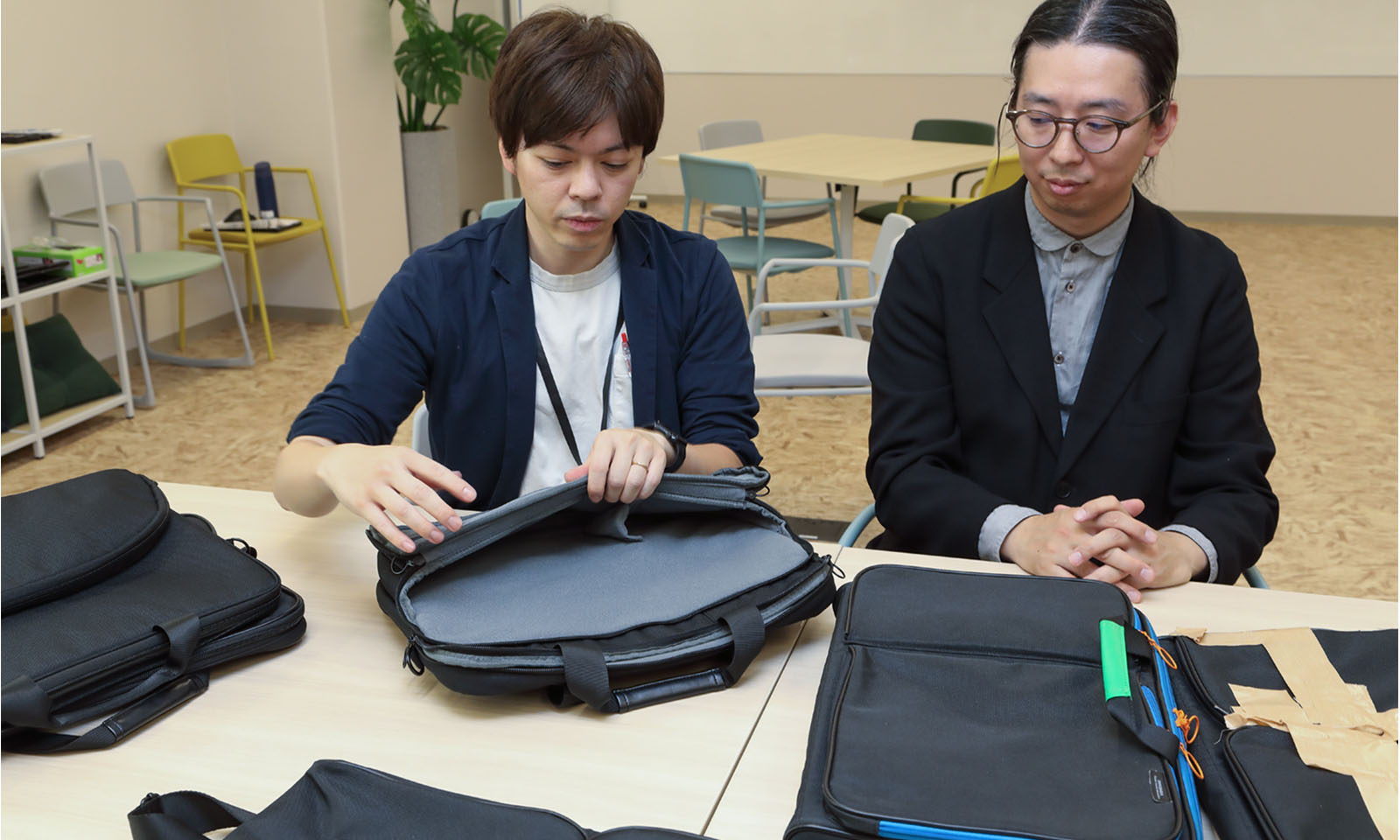 ALTはじまり。黒いジャケットを羽織った男性2人が並んで座っている。左側の男性は、パソコンバッグを手元に置いて、左手で外側のポケット中央付近をつかんで大きく開き、右手でファスナーのあたりをなぞっている。右側のメガネをかけた男性は、両手を机の上で組んでその様子をじっと見ている。ALTおわり。