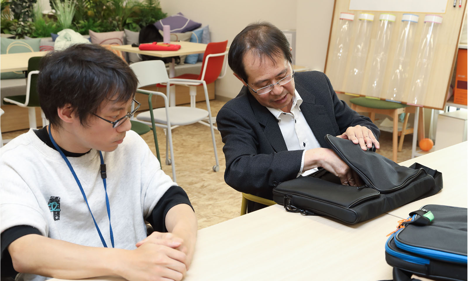 ALTはじまり。メガネをかけた2人の男性が並んで座っている。黒いジャケットを羽織った右側の男性は、黒いパソコンバッグの外側に2つ付いているポケットの片方を開いて左手で押さえ、右手で物を出し入れする様子を再現している。左側の白い服の男性は、その様子を真剣な面持ちで見守っている。ALTおわり。