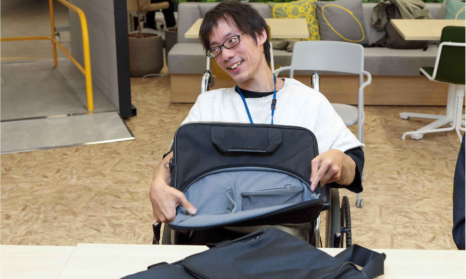 ALTはじまり。メガネをかけた車いすに乗った男性が、自分の膝の上にパソコンバッグを立てて置き、外側に付いているラウンド形状のポケットを右手で押さえて手前側に大きく広げている。向かって左側に視線を送りながら、にこやかな表情で話している。ALTおわり。