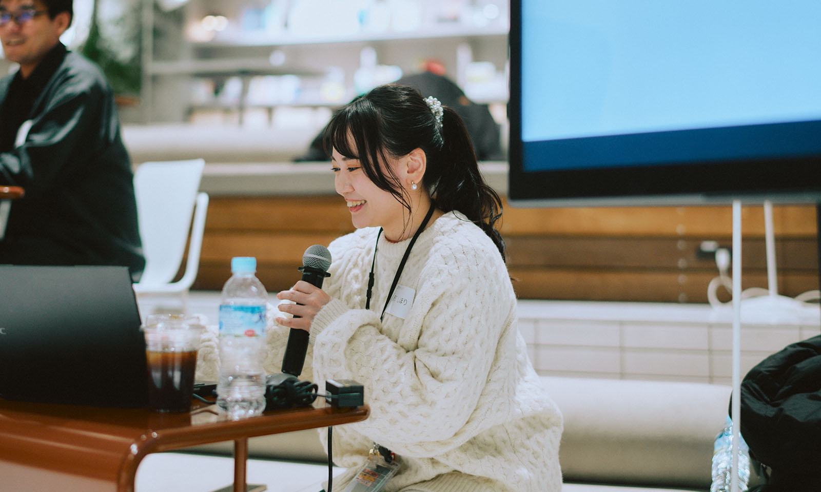 ALT：ALTはじまり。白いセーターを着て、白で合わせたヘアアクセサリーを使い長い髪を高い位置でひとつに結んでいる野口さん。ディスプレイの横で、座りながらマイクを持って登壇者に質問をしている。ALTおわり。
