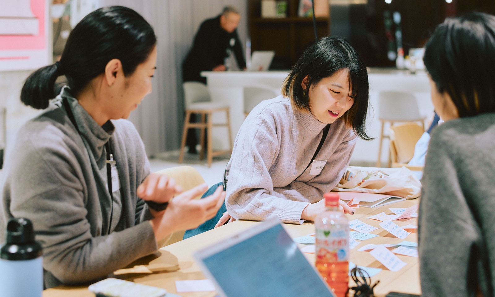 ALTはじまり。会場全体の様子。広々としたスペースの奥には白で統一された棚が並んでいる。その手前にベンチタイプのソファが1列並び、その手前に設置された椅子に登壇者の2人が腰掛けている。さらに手前に、参加者が20名ほど、明るいナチュラルなベージュ色で揃えられたテーブルと椅子のスペースに腰掛けている。参加者の年齢、性別は様々で、話をする田中さんの姿に視線を向けている。ALTおわり。