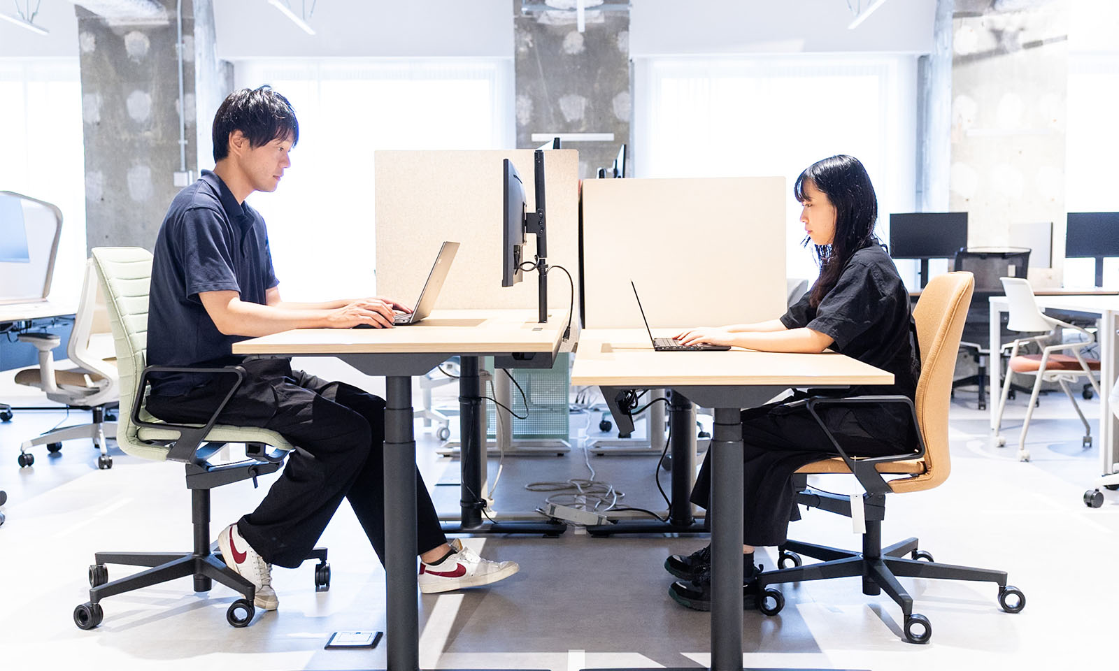 ALTはじまり。画像：昇降デスクが2台向かい合って並べられていて、背の高い男性と背の低い女性がそれぞれ自分の身長に合わせた高さに調整してパソコンに向かって作業をしている。身長に合わせて高さを調整できるのも昇降デスクの基本価値のひとつ。画像おわり。ALTおわり。